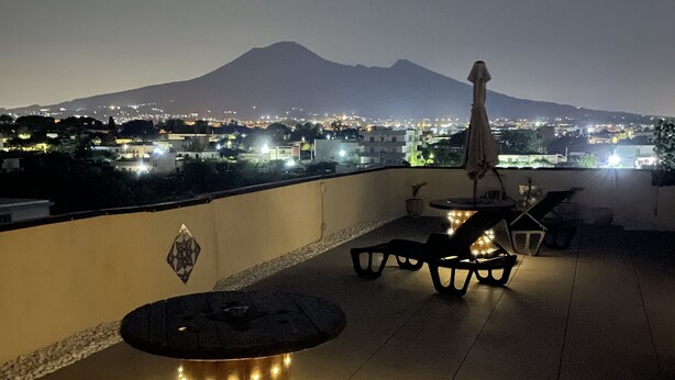 A rooftop terrace with a round table and chairs, overlooking a cityscape with a mountain in the background. - Pompei Window (Pompei)