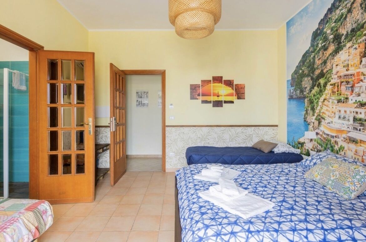 A bedroom with a bed, a door, a wall with a sunset painting, and a mural of a coastal town.