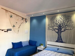 Une chambre à coucher avec un lit, deux canapés bleus, une table de chevet avec un vase et une tapisserie murale représentant un arbre.