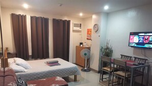 A hotel room with a bed, a sofa, a dining table with chairs, a TV, and a fan. - ANGEL ROOM HOMESTAY AT KASSEL RESIDENCES (Parañaque)