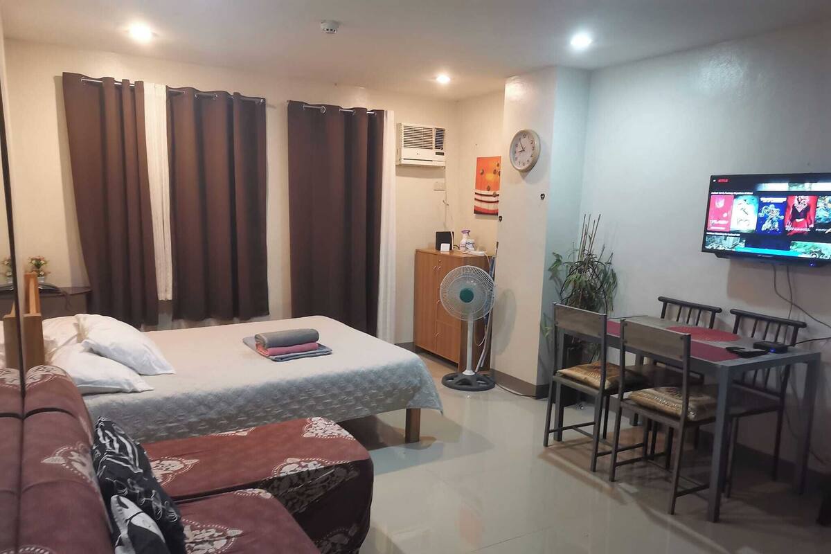 A hotel room with a bed, a sofa, a dining table with chairs, a TV, and a fan.