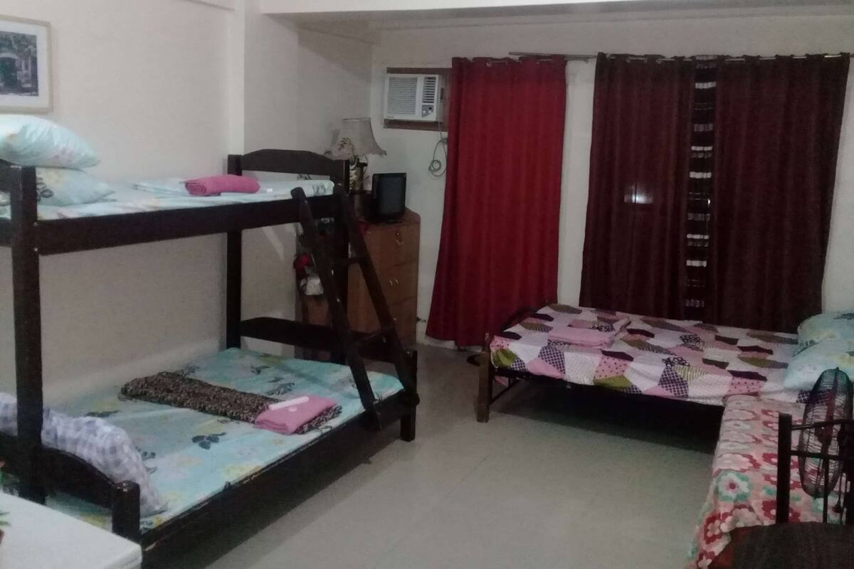 A dormitory room with bunk beds, a desk with a chair, a window with curtains, and an air conditioning unit.