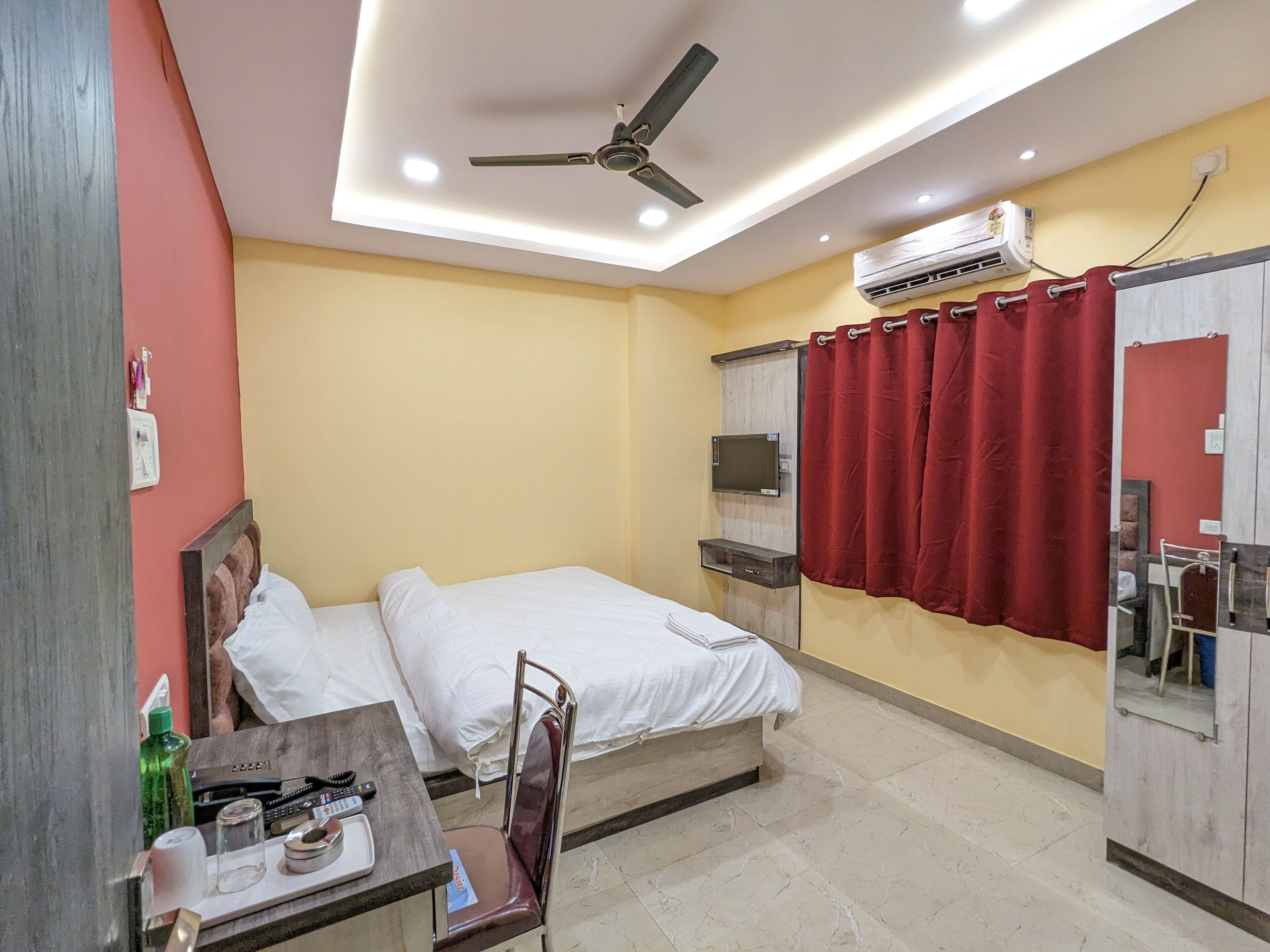 Une chambre d’hôtel avec deux lits, un bureau avec une chaise, un ventilateur de plafond, un appareil de climatisation et un rideau rouge.