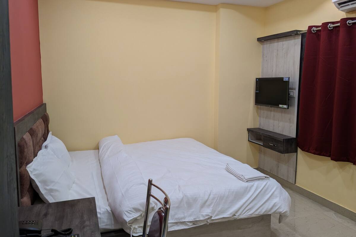 A neatly made bed in a room with a television mounted on the wall, a red curtain, and a wooden headboard.