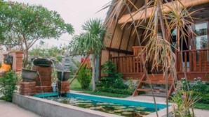Edificio rústico con techo de paja, rodeado de un jardín con un pequeño estanque y diversas plantas.