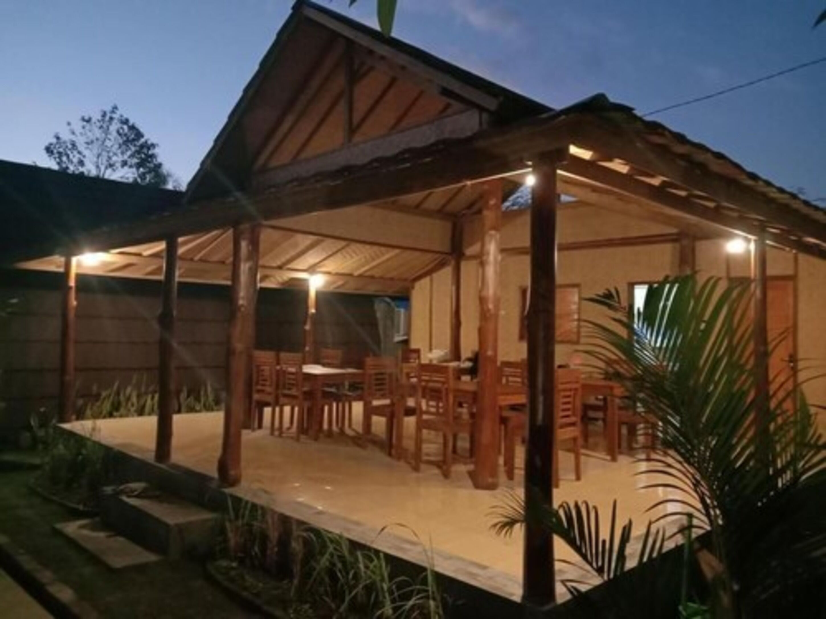 Un área de comedor al aire libre con mesas y sillas de madera, rodeada de plantas y un techo cubierto.