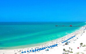 A beach with clear turquoise waters, a long pier extending into the sea, and numerous blue umbrellas lined up along the shore. - Soothing Spa, Patio, Games & Grill @ Mystic Haven (Largo)