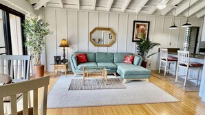 A living room with a sectional sofa, a coffee table, a side table with a lamp, a mirror, and a dining area with chairs. - Tranquility on West Moloka'i (Maunaloa)