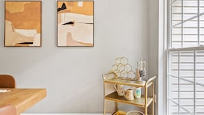 A room with a wooden dining table, two abstract paintings on the wall, a gold metal shelf with decorative items, and a window with white blinds. - NEW! 3 Bdrm | Mt Washington | Garage (Pittsburgh)