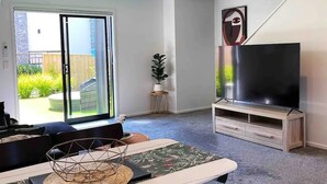 Ein modernes Wohnzimmer mit großem Flachbildfernseher auf Holzfuß, einem Essbereich mit Tisch und Stühlen und einer Schiebeglastür zum Garten.