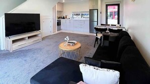 Ein modernes Wohnzimmer mit Flachbildfernseher, schwarzem Sofa und Essbereich mit Tisch und Stühlen.