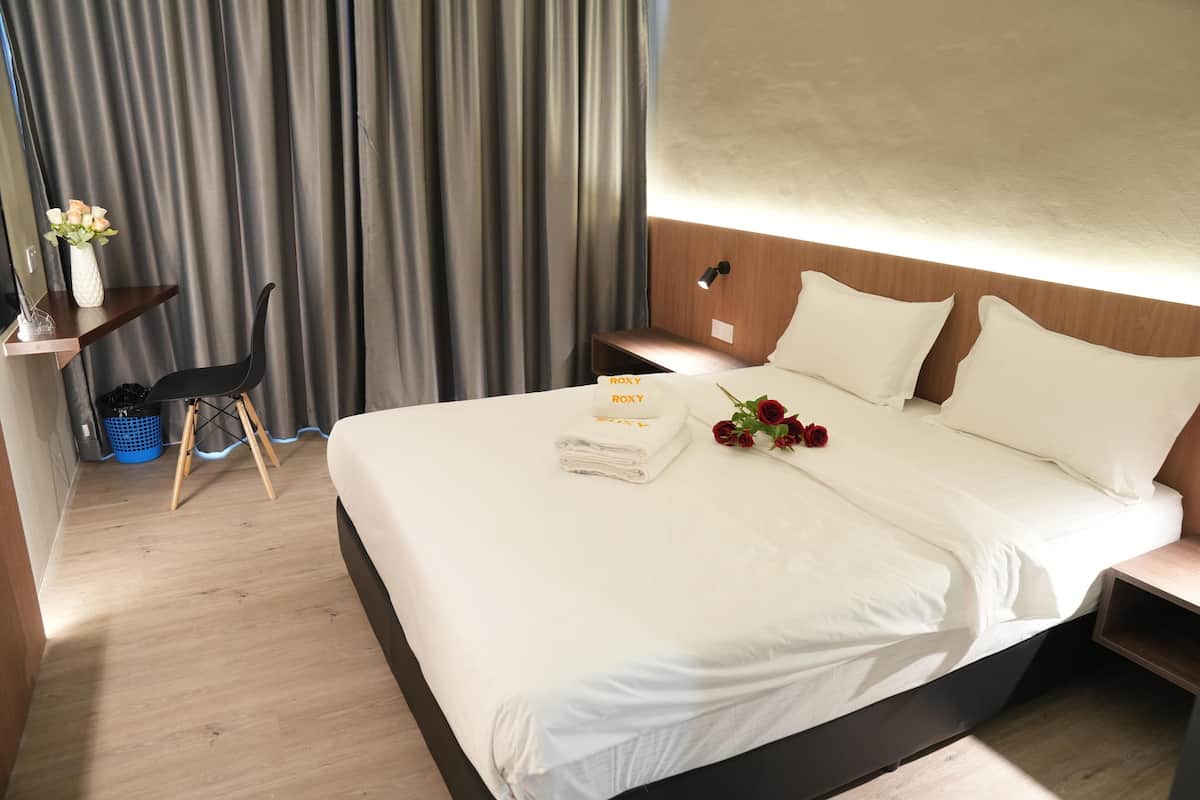 A hotel room with a neatly made bed, a bedside table with a lamp, a small desk with a chair, and a window with curtains.
