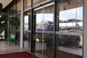 Entrada moderna de un edificio con puertas de vidrio, un banco verde y el reflejo de un coche y una calle en el cristal.