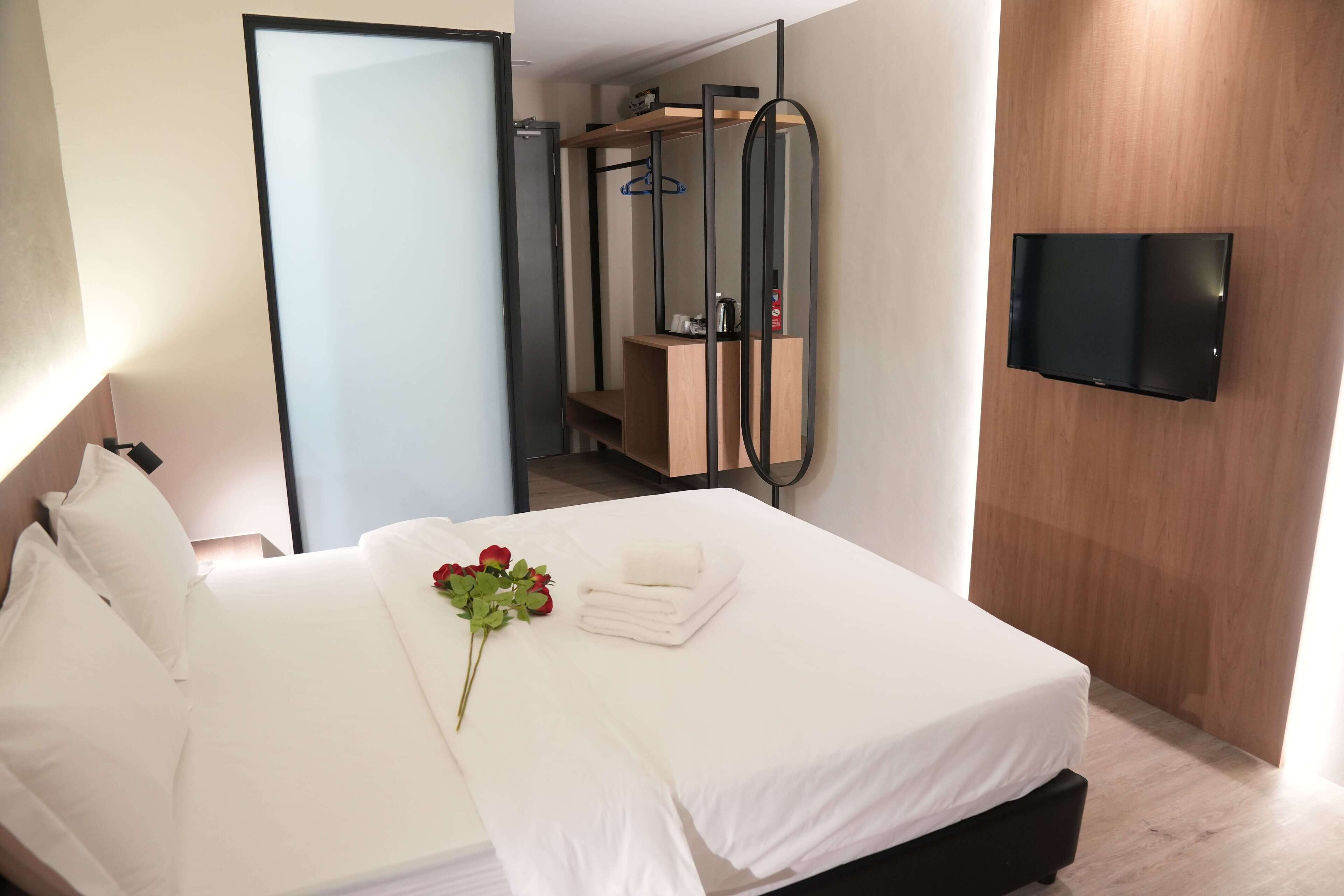 A hotel room with a neatly made bed, a wooden wardrobe, a mirror, and a flat-screen TV mounted on the wall.