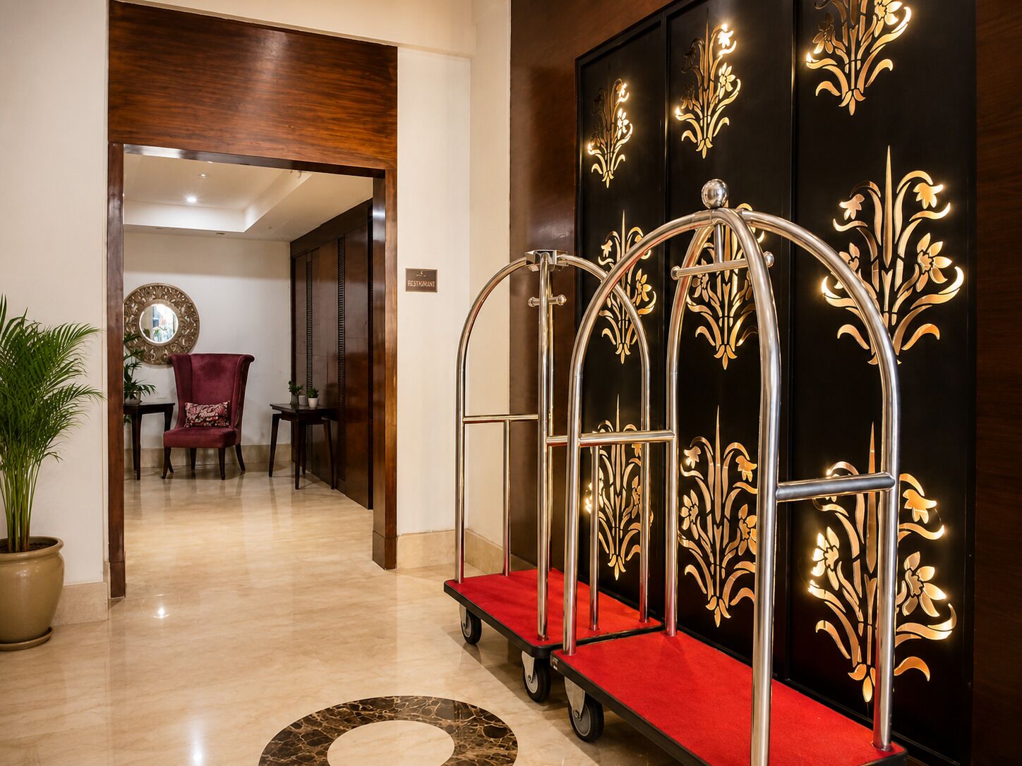 Un hall d’hôtel avec un tapis rouge, un chariot en métal et un mur orné de motifs floraux dorés.