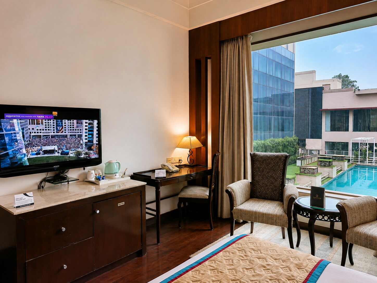 A hotel room with a television, desk, chairs, a bed, and a view of a swimming pool.