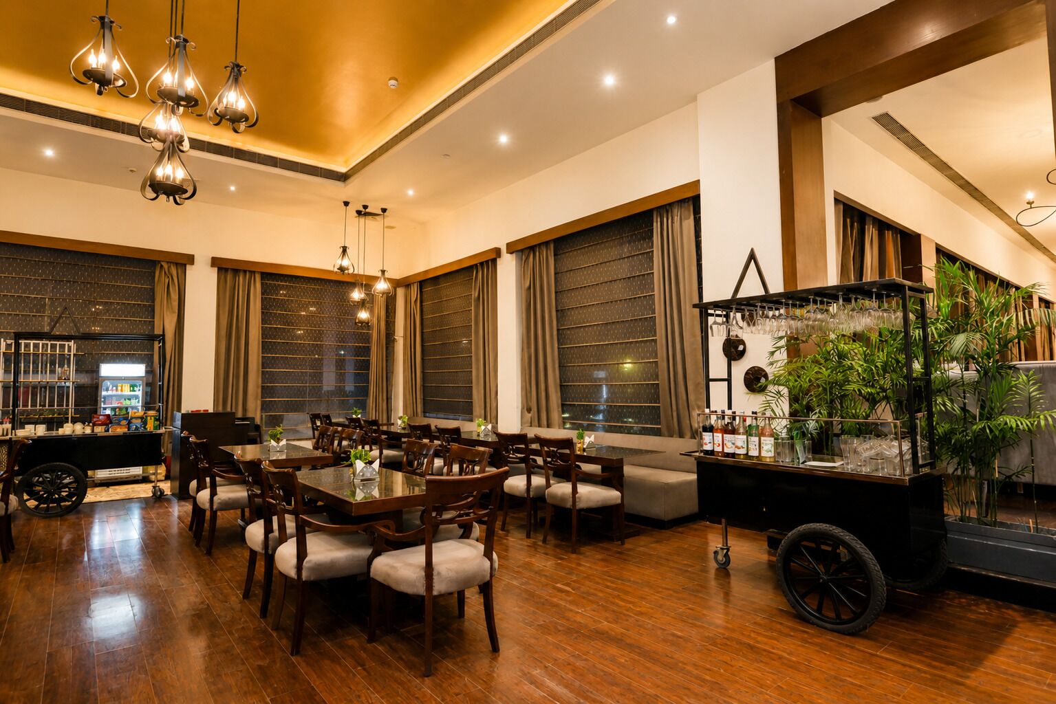 A spacious dining area with wooden tables and chairs, large windows with curtains, and a bar area with a plant display.