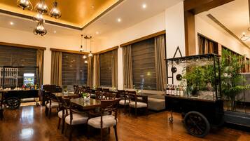 A spacious dining area with wooden tables and chairs, large windows with curtains, and a bar area with a plant display.