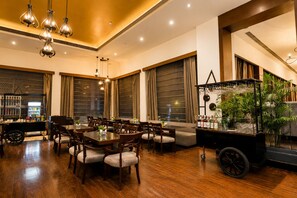 Un amplio comedor con mesas y sillas de madera, ventanas grandes con cortinas y una zona de bar con un arreglo de plantas.