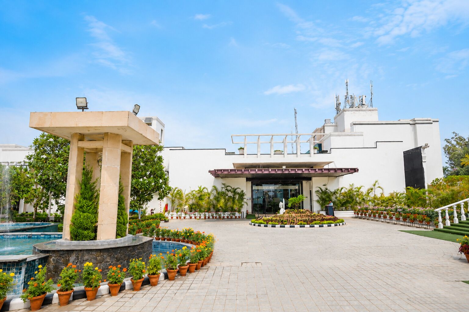A hotel entrance with a pool, potted plants, and a clear sky.