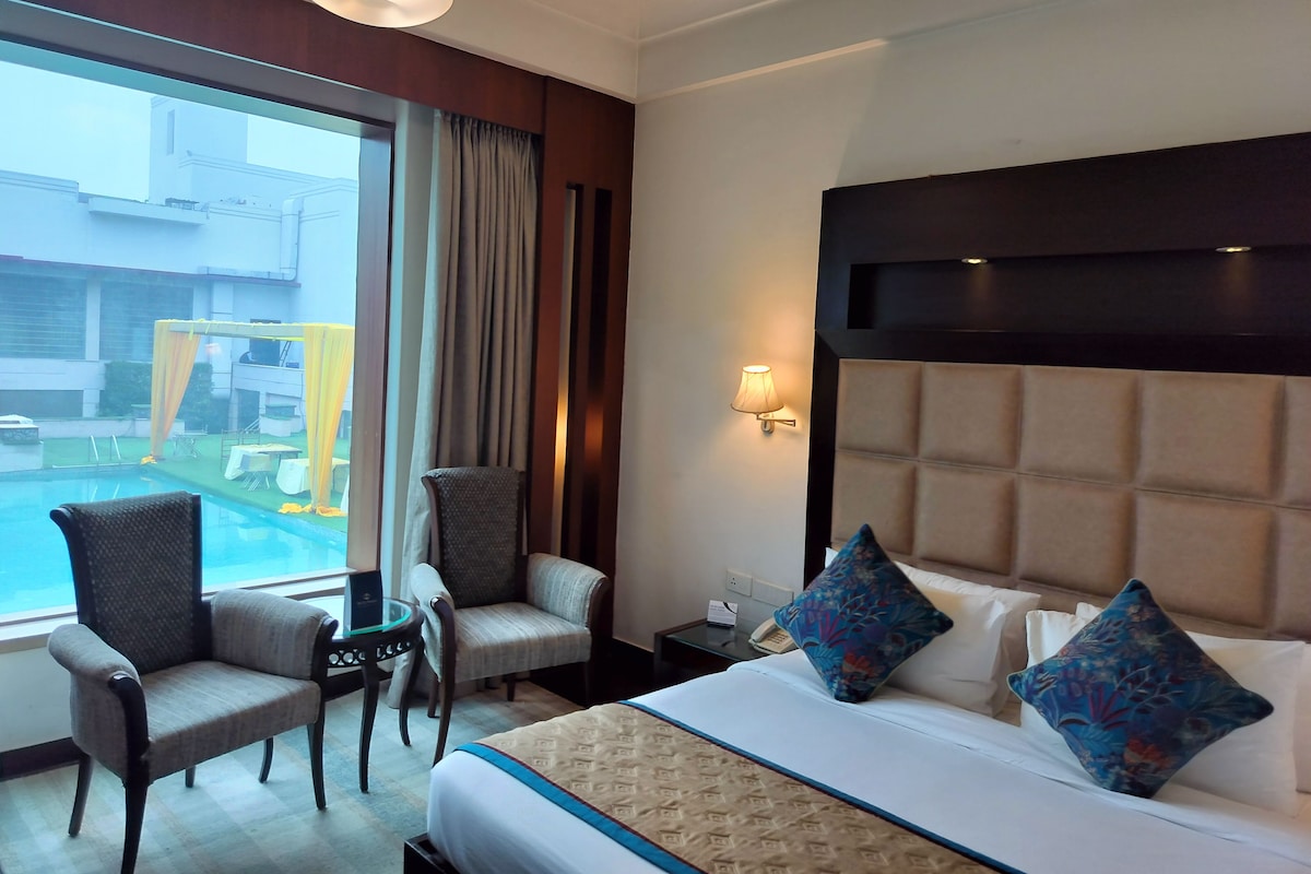 A hotel room with a bed, two chairs, a small table, a nightstand, a lamp, and a view of the outside.