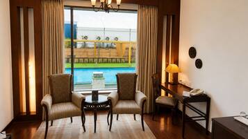 Habitación de hotel con cama, dos sillones, una mesita, un escritorio con lámpara y vista a una piscina.