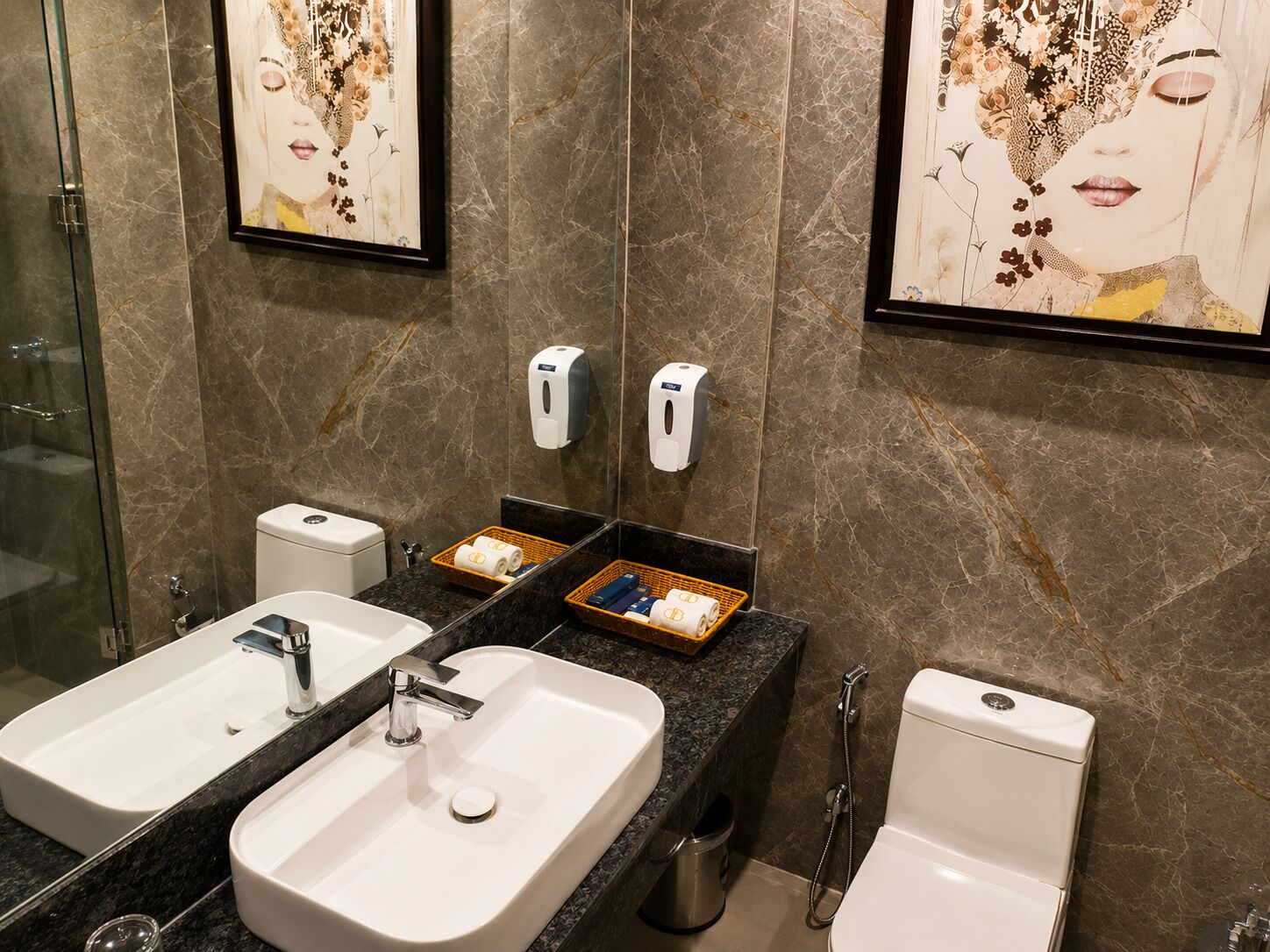 A modern bathroom with two sinks, a toilet, and a shower. There are hand dryers and a soap dispenser on the wall.