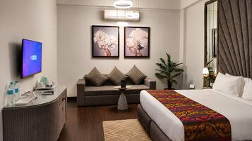 A hotel room with a bed, a sofa, a desk, a TV, and artwork on the wall.