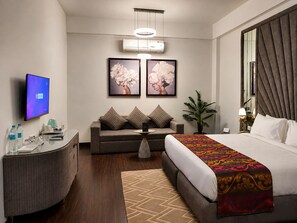 A hotel room with a bed, a sofa, a desk, a TV, and artwork on the wall.