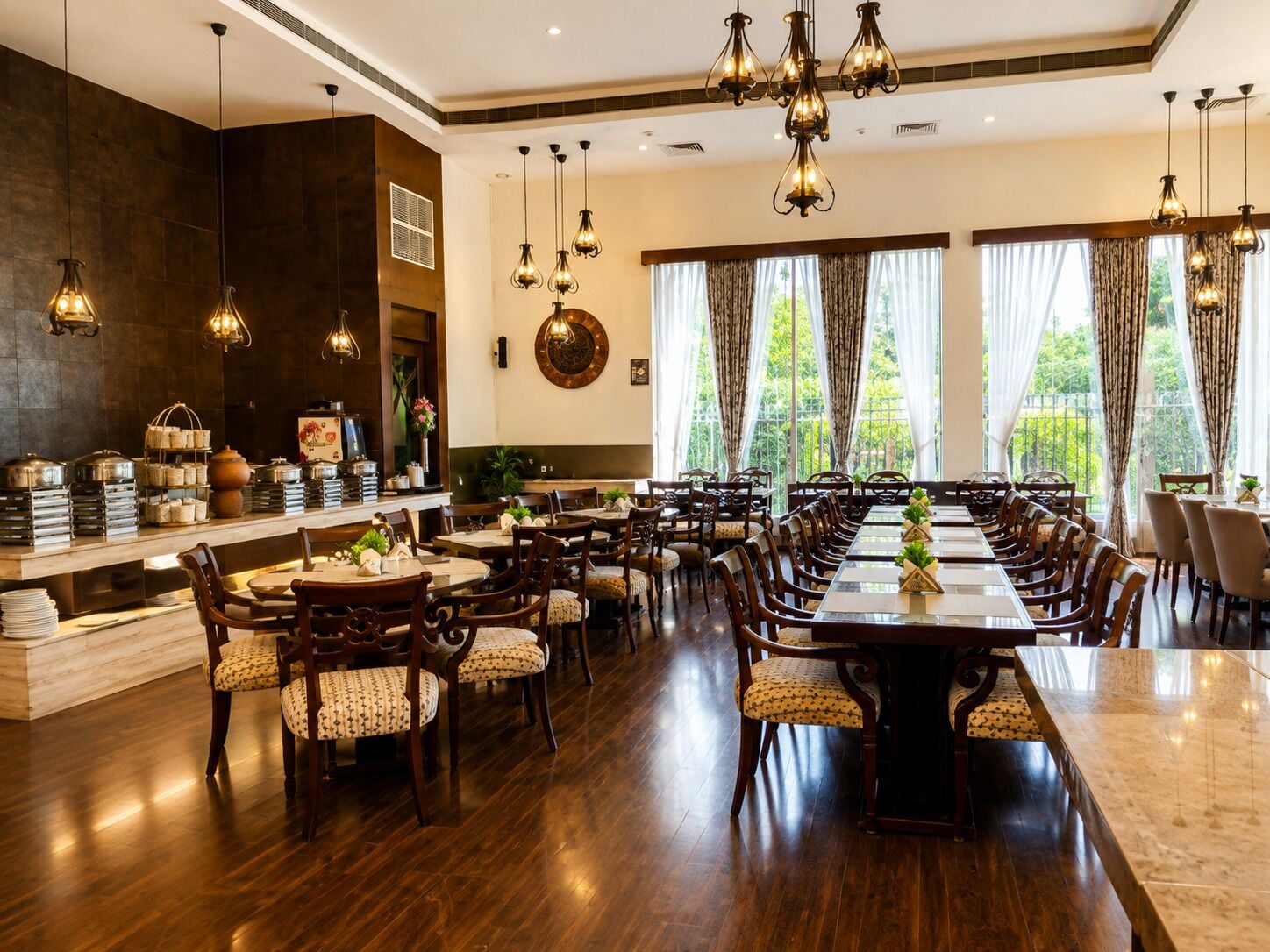 A spacious dining area with multiple tables and chairs, a buffet setup on the left, and large windows with curtains.