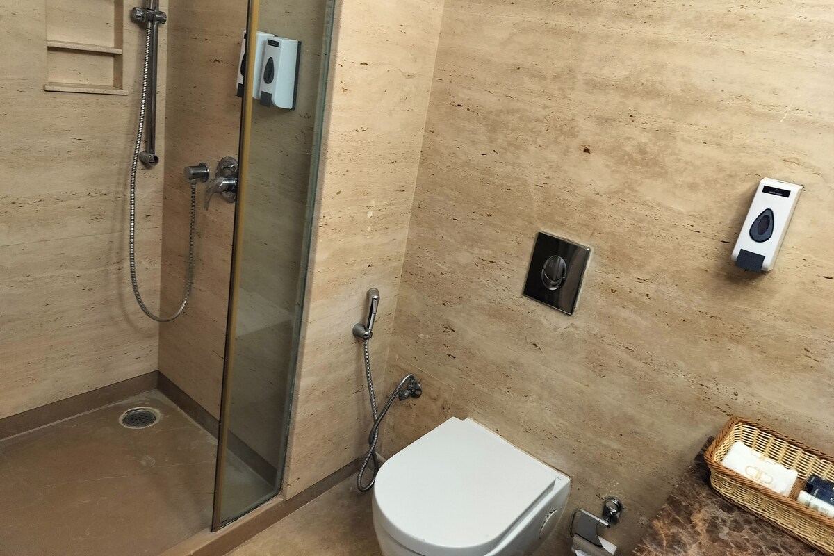 Un baño con ducha envuelta en vidrio, inodoro y secamanos fijado a la pared.
