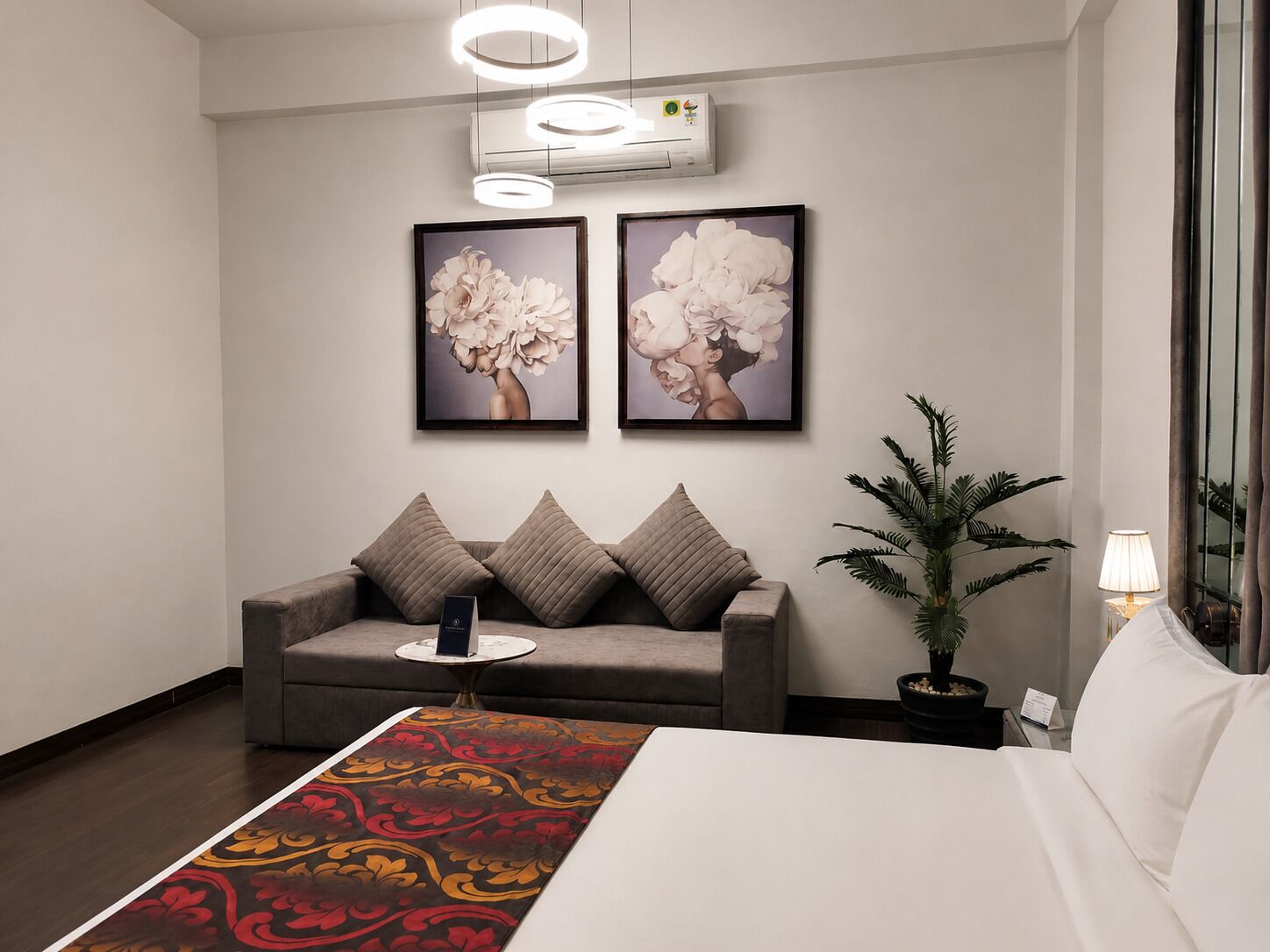 A hotel room with a bed, a sofa, a coffee table, a potted plant, and two framed artworks on the wall.