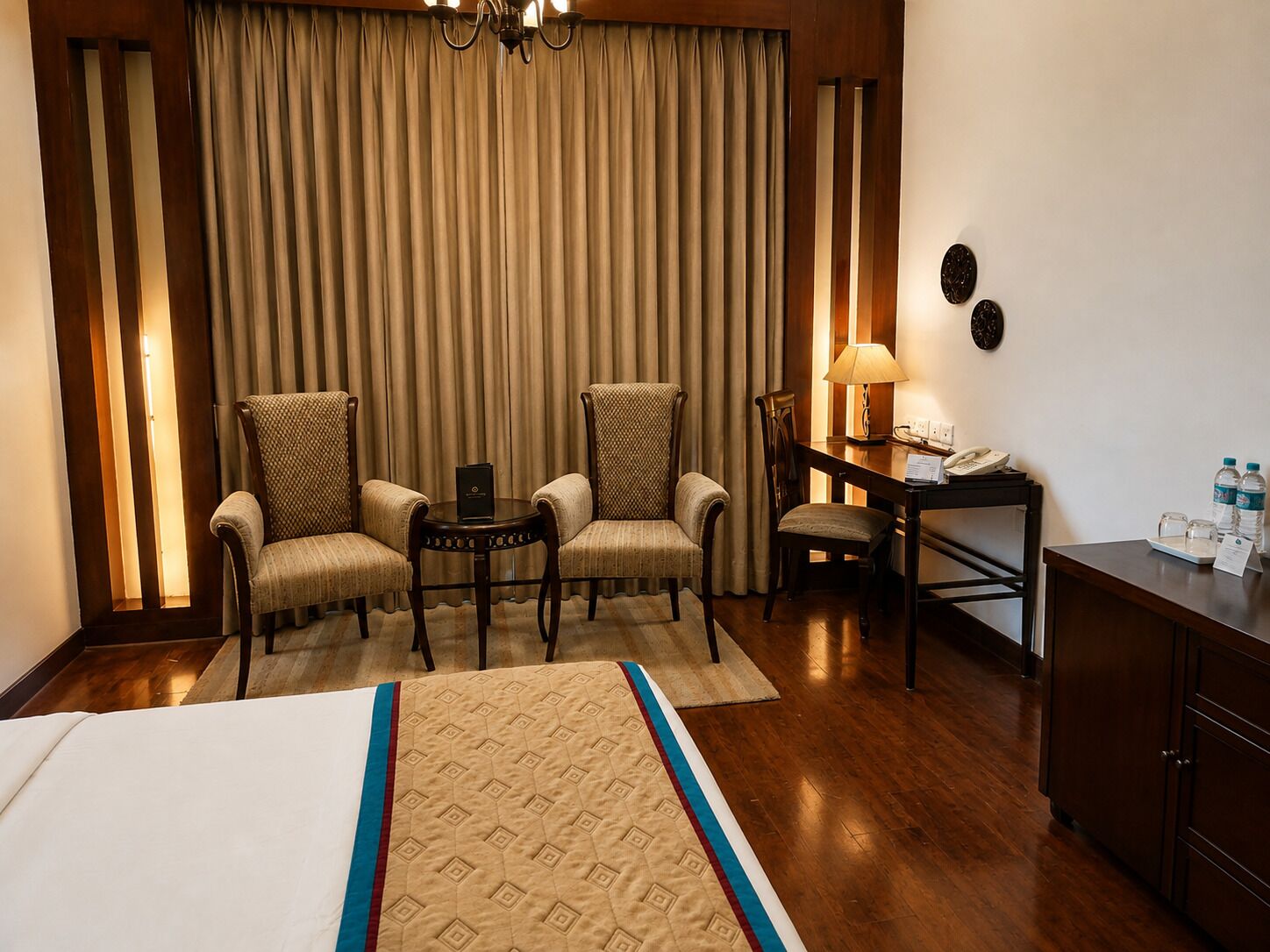 Habitación de hotel con cama, dos sillones, un escritorio con lámpara, teléfono y un armario con botellas de agua.