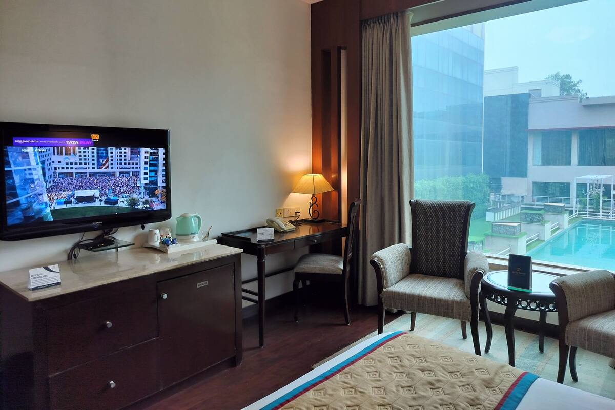 A hotel room with a bed, a desk with a lamp, a television, and a view of a pool and buildings.