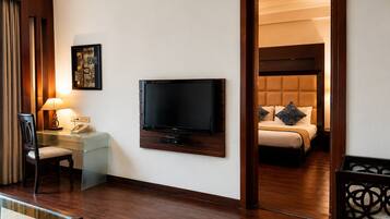 A hotel room with a flat-screen TV mounted on the wall, a bedside table with a lamp, a chair, and a bed with a headboard and pillows.