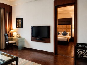 Habitación de hotel con televisor de pantalla plana montado en la pared, mesita de noche con lámpara, una silla y cama con cabecera y almohadas.