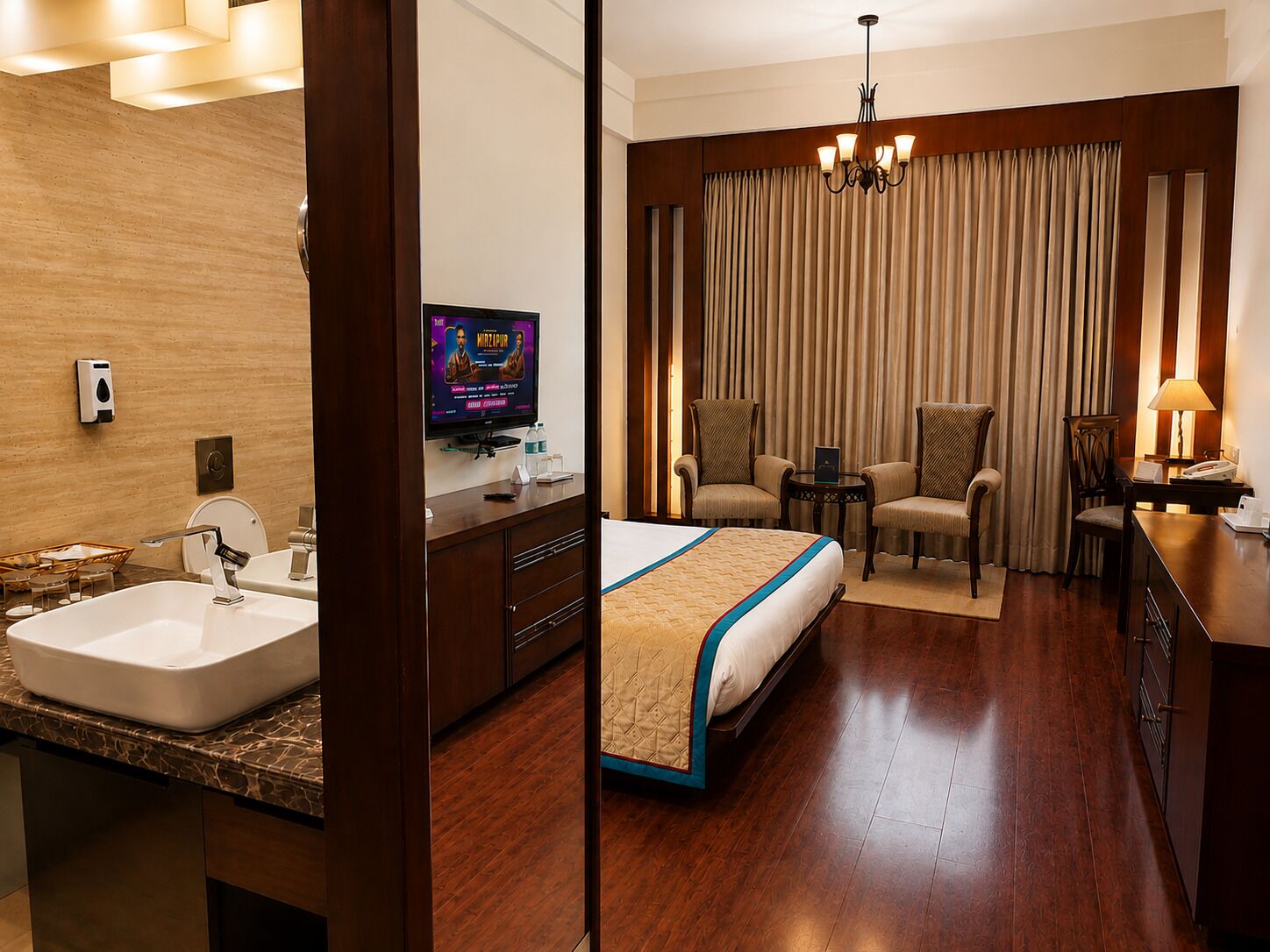 Habitación de hotel con cama, escritorio, silla, televisor y baño con lavamanos y espejo.