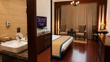 Habitación de hotel con cama, escritorio, silla, televisor y baño con lavamanos y espejo.