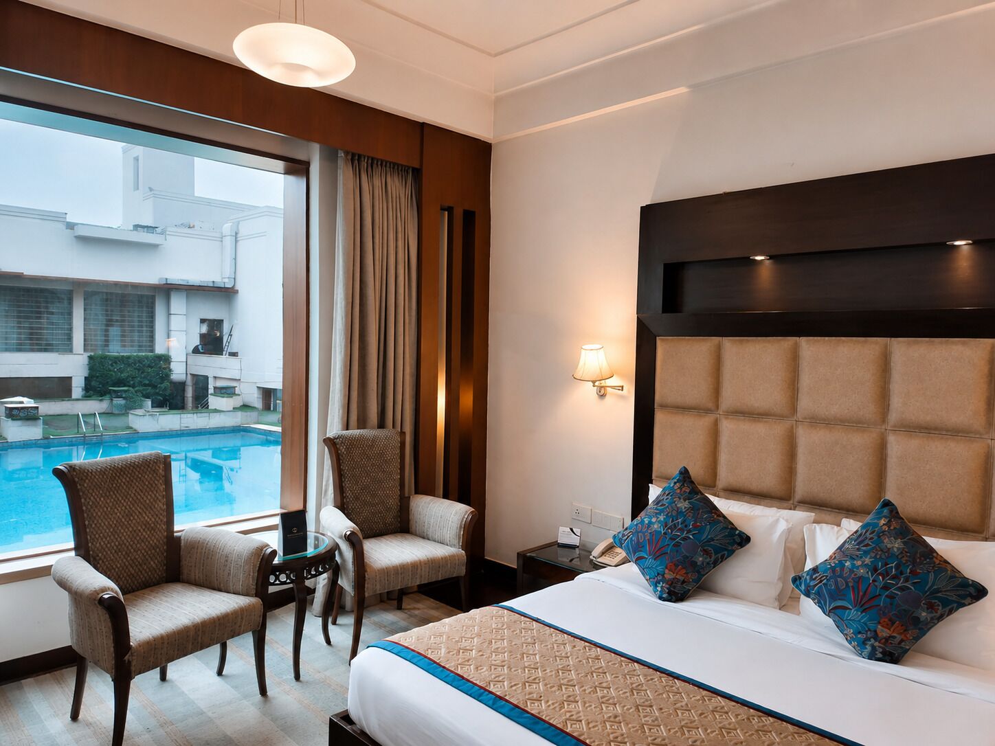 A hotel room with a large bed, two chairs, a small table, a bedside lamp, and a view of a swimming pool.