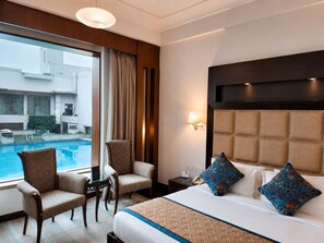 A hotel room with a large bed, two chairs, a small table, a bedside lamp, and a view of a swimming pool. - Bel-La Monde Hotel NH8 at 6km from IGI Airport Delhi (New Delhi)