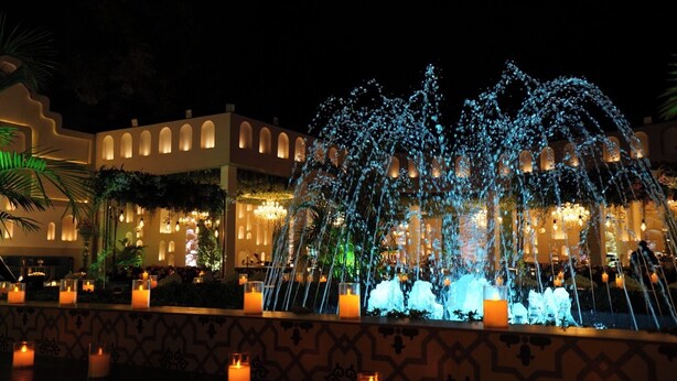 A nighttime view of a hotel with a central water fountain, candle-lit tables, and a patterned wall.