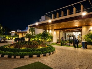 Entrada de hotel con un paseo techado, palmeras y un estacionamiento.