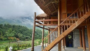 Ein hölzernes Gebäude mit einer Treppe, die zu einem Balkon führt, umgeben von üppiger Vegetation und einem bewölkten Himmel.