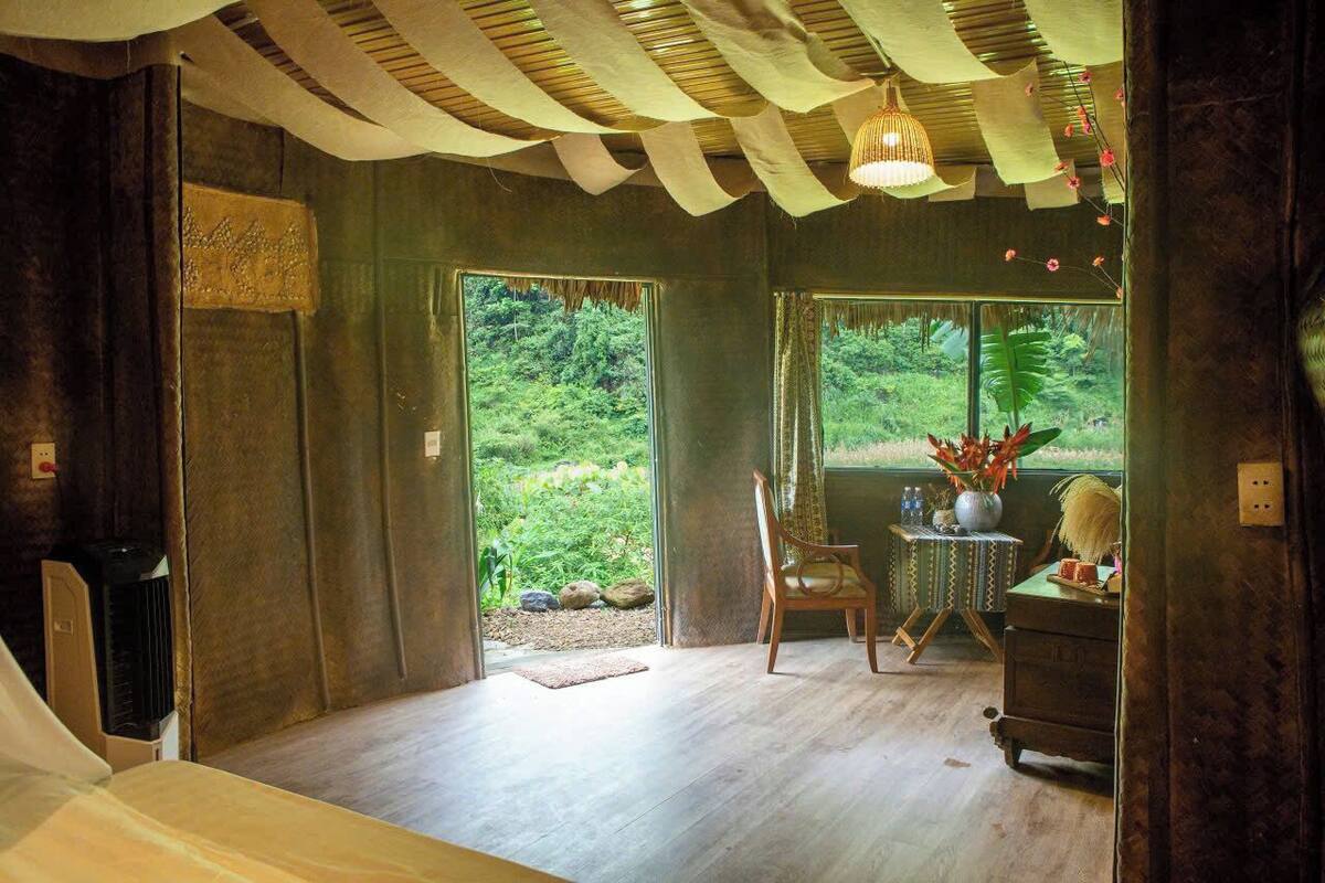 A room with a wooden floor, a chair, a small table with a vase, and a window with a view of greenery.