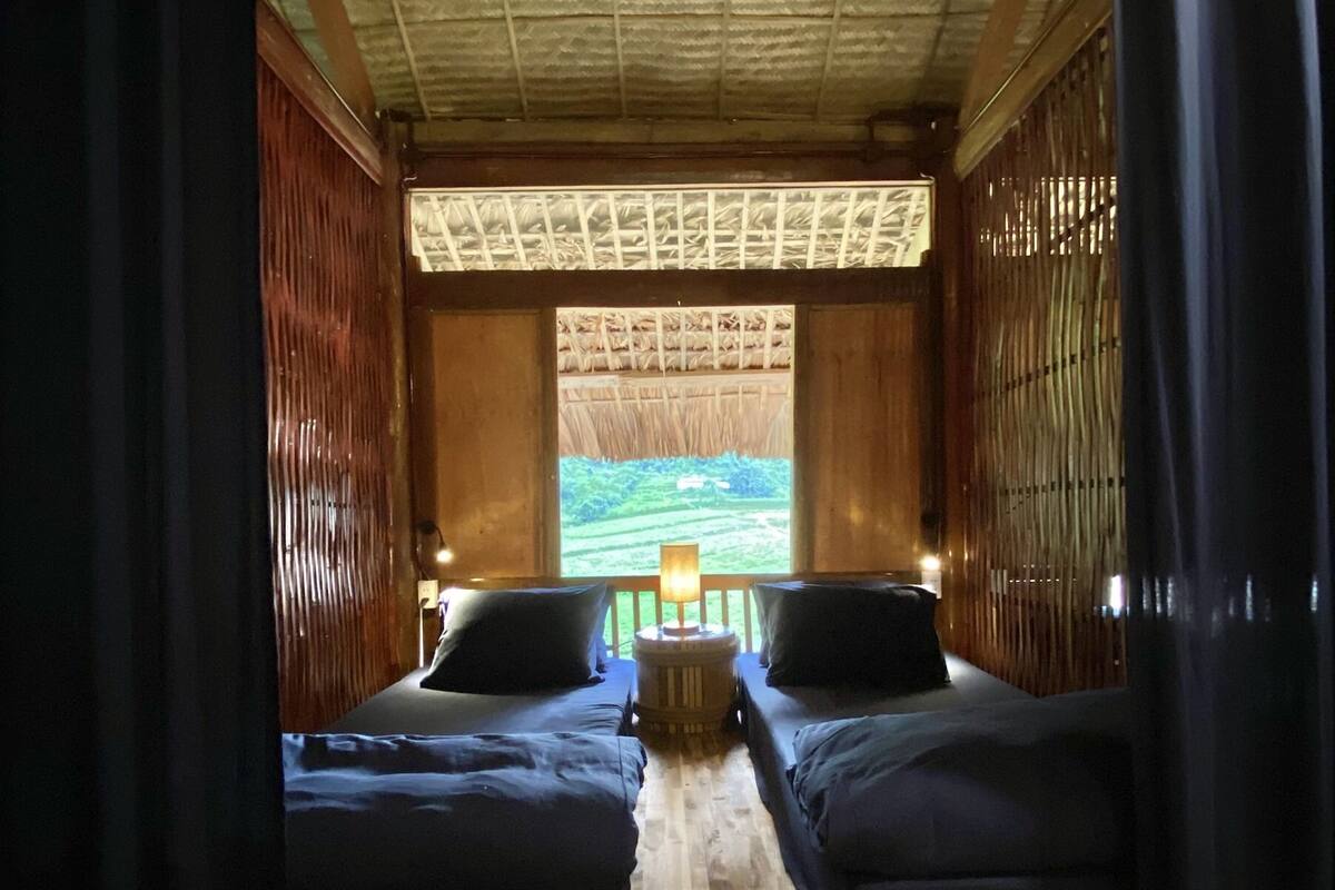 A room with two beds, wooden flooring, a window with a view of greenery, and a ceiling with a patterned design.