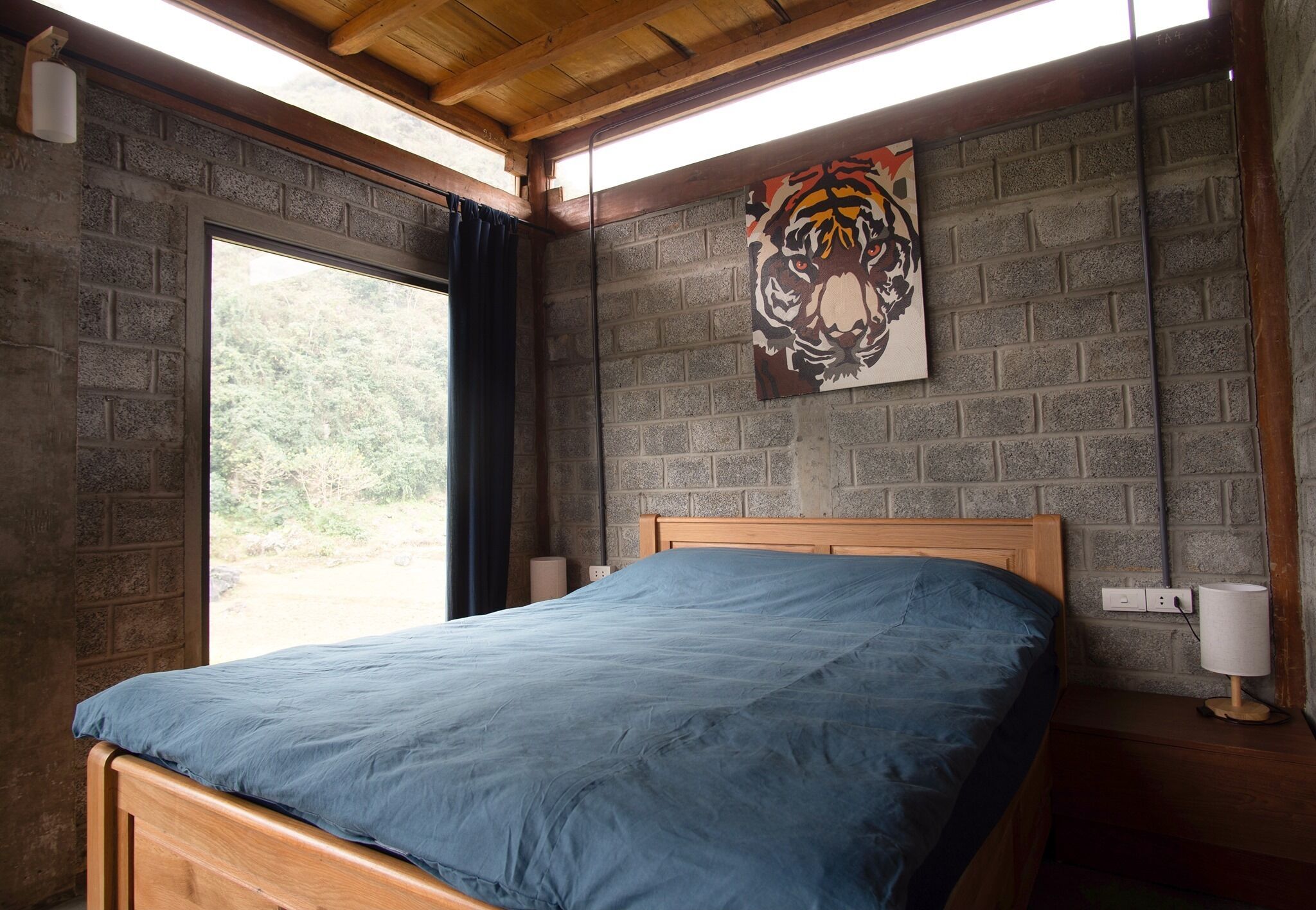 A bedroom with a wooden bed, a brick wall, a tiger painting, and a window with a view of trees.