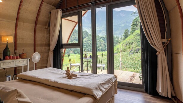 Ein Schlafzimmer mit einem Bett, einem Nachttisch mit Lampe, einem Ventilator und Blick auf eine Berglandschaft durch große Fenster.