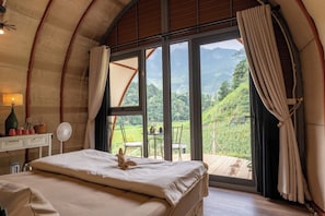A bedroom with a bed, a bedside table with a lamp, a fan, and a view of a mountainous landscape through large windows. - To Day Du Gia Village (Du Gia)