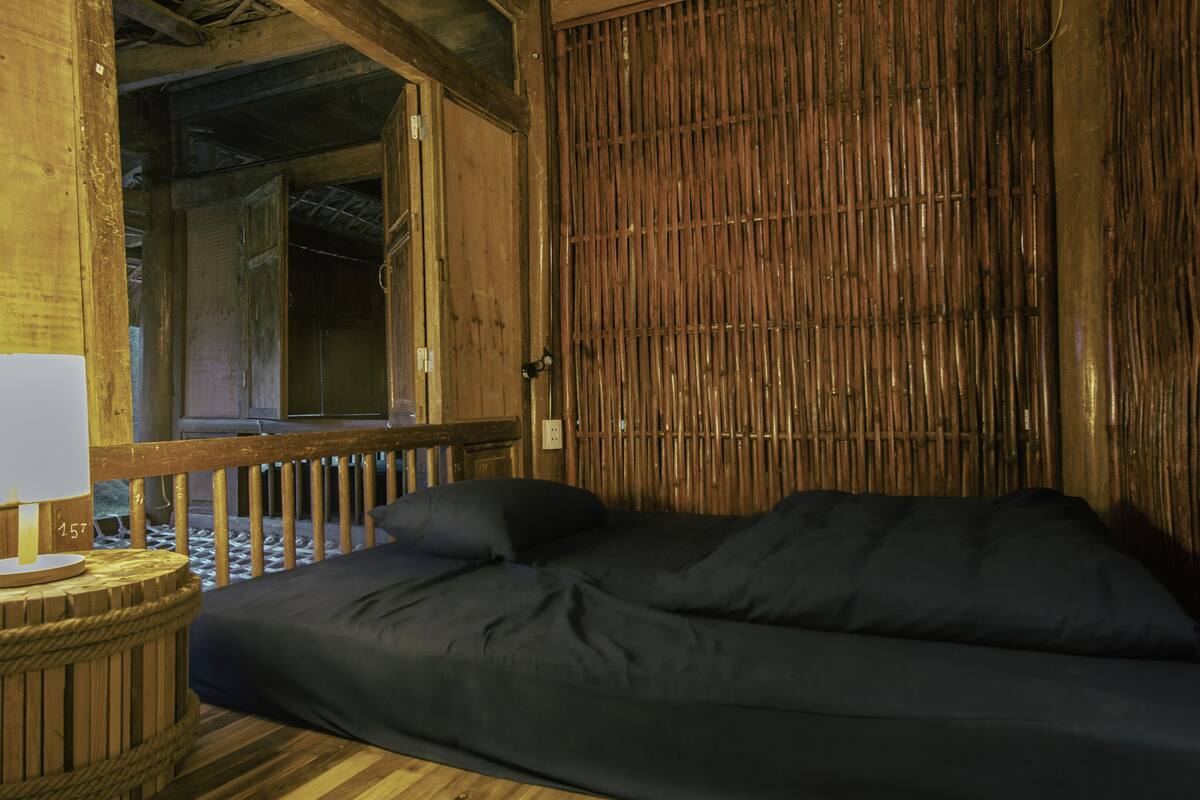 A room with a wooden floor, a bed with a dark cover, a lamp on a wooden table, and a bamboo wall.