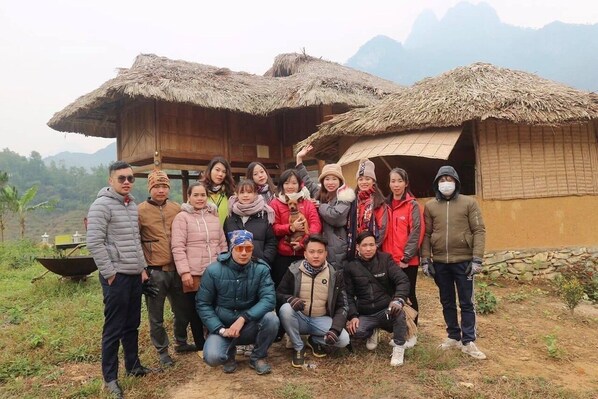 A group of people posing in front of a traditional thatched-roof hut with a dirt ground and mountains in the background. - To Day Du Gia Village (Du Gia)